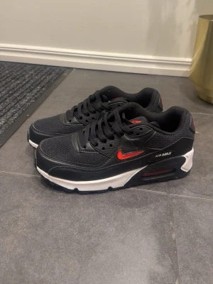 Nike Air Max 90 svart/röd sneakers - Snygga Nike Air Max 90 sneakers i svart med röda detaljer och vit sula. Skorna har en klassisk siluett med mesh och läderpaneler, samt den ikoniska Air Max-dämpningen i sulan. Perfekta för dig som gillar streetwear och sportig stil.