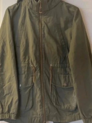 Olivgrön parkasjacka med huva - Snygg olivgrön parkasjacka med dragkedja framtill, justerbar midja med snören och två stora fickor med lock och knapp. Jackan har huva och resår vid ärmsluten. Perfekt för blåsiga dagar och enkel att matcha till olika outfits. Liten i storlek är en 134