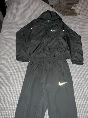 nike miller - Snyggt svart träningsset från Nike med jacka och byxor. Jackan har huva, dragkedja och två fickor, samt reflekterande Nike-logga. Byxorna har resår i midjan och matchande logga. Perfekt för löpning eller träning. Materialet är lätt och andas. Passar 160-170cm