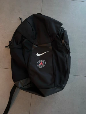 Svart PSG ryggsäck med regnskydd  - Snygg svart ryggsäck från Nike med PSG-logga på framsidan och vit Nike Swoosh. Ryggsäcken har flera fack med dragkedja och är tillverkad i slitstarkt syntetmaterial. Perfekt för dig som gillar sportig stil och vill visa ditt stöd för Paris Saint-Germain.