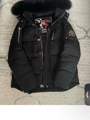Svart dunjacka från Moose Knuckles - Snygg svart dunjacka från Moose Knuckles med pälskantad huva och silverfärgade dragkedjor. Jackan har logotyp på ärmen, flera fickor och quiltad design. Perfekt för kalla vinterdagar och riktigt varm.