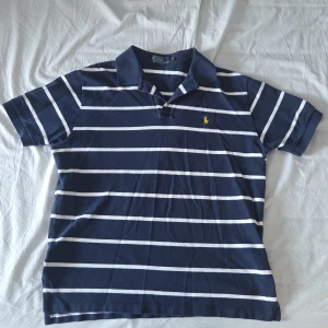 Marinblå randig pikétröja Polo Ralph Lauren - Klassisk marinblå pikétröja från Polo Ralph Lauren med vita horisontella ränder och gul broderad logga på bröstet. Tröjan har krage, korta ärmar och knäppning vid halsen. Perfekt för en avslappnad men stilren look.