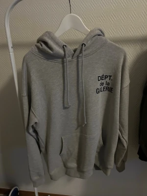 Grå hoodie DÉPT. de la GALERIE - Snygg grå hoodie från DÉPT. de la GALERIE med svart tryck både fram och bak. Klassisk känguruficka, justerbar huva med snören och ribbade muddar. Perfekt för en chill och trendig look. Tillverkad i mjukt material för extra komfort. Äkta men lågt pris vill bara bli av med den