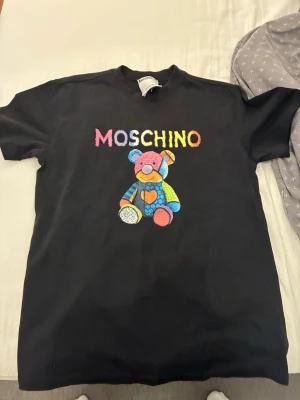 Svart Moschino t-shirt med nalle - Cool svart t-shirt från Moschino med färgglad nalle och logga framtill. T-shirten har rund hals och korta ärmar. Perfekt statement-plagg för dig som gillar lekfull design och streetstyle.