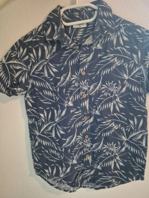  Barn Blå kortärmad skjorta med palmblad stl 134 - Snygg blå skjorta från Twill med korta ärmar och vita palmbladsmönster. Skjortan har klassisk krage och träfärgade knappar framtill. Perfekt för sommarens alla äventyr och ger en chill vibe.