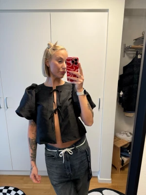 Svart blus med knyt från ginatricot - Trendig svart blus från Gina med korta puffärmar och två knyt framtill. Croppad modell som ger en cool och modern look. Perfekt att styla med höga jeans eller kjol.