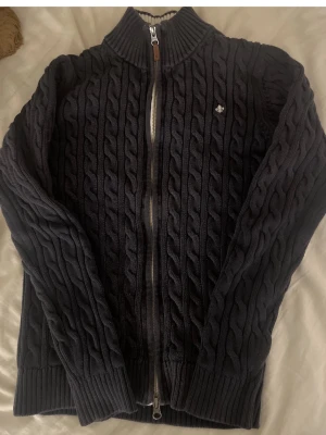 Mörkblå Morris tröja - Kabelstickad mörkblå tröja med dragkedja framtill och hög krage. Tröjan har ribbade muddar och en liten broderad logga på bröstet. Perfekt för lager på lager och en snygg casual stil.