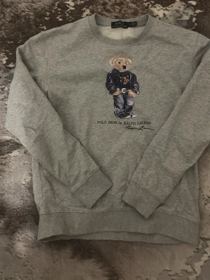Grå sweatshirt Polo Bear Ralph Lauren - Grå sweatshirt från Polo Ralph Lauren med ikoniskt Polo Bear-motiv på bröstet. Rund halsringning, ribbade muddar vid ärmslut och nederkant. Perfekt för en avslappnad och trendig look.