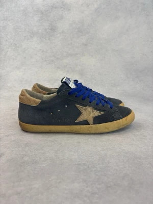 Golden Goose sneakers med blå snören - Mörkgrå sneakers från Golden Goose med beige stjärndetalj på sidan, blå snören och beige häl. Ovandelen är i mocka och sulan har en vintage-look. Klassisk låg modell med GGDB-logga på plösen.