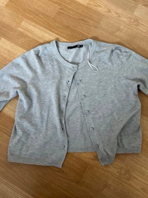 Ljusgrå kofta - Ljusgrå kofta med rund halsringning och knäppning framtill. Koftan har långa ärmar och ribbade muddar vid ärmslut och nederkant. Perfekt att slänga över en t-shirt eller linne för en chill look.