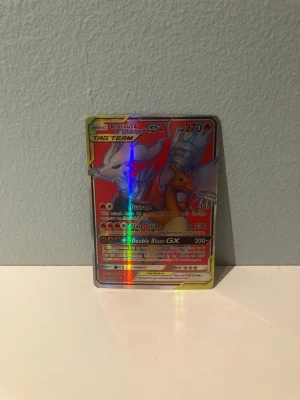 Pokémon kort Reshiram & Charizard GX - Säljer ett holografiskt Pokémonkort med Reshiram & Charizard GX från TAG TEAM-serien. Kortet har färgstark design med röd, gul och blå bakgrund och tydliga detaljer. Perfekt för samlare eller Pokémonfans som vill utöka sin samling med ett coolt och unikt kort.