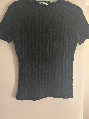 Svart ribbad topp från Zara - En svart ribbad topp från Zara med korta ärmar och rund halsringning. Toppen har en åtsittande passform och ett vågigt avslut vid hals och nederkant. Perfekt att styla med jeans eller kjol. 