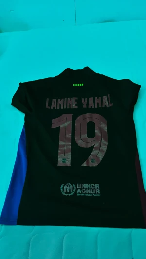 FC Barcelona Lamine Yamal matchtröja - Säljer en svart/blå FC Barcelona matchtröja med Lamine Yamal och nummer 19 på ryggen. Tröjan har klubbmärke och Spotify-logga på bröstet samt UNHCR-tryck nedtill. Sidorna har vinröda och blå paneler. Perfekt för fotbollsfans!