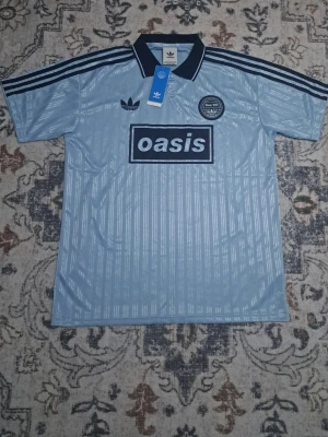 Ljusblå Adidas Oasis tröja - Säljer en ljusblå retroinspirerad Adidas tröja med svart krage och svarta ränder på axlarna. Framtill finns ett stort Oasis-tryck och ett Live Forever 25-märke. Tillverkad i glansigt, lätt material och har klassisk Adidas-logga på bröstet. Perfekt för dig som gillar sportig stil.live oasis tröja