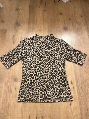 Leopardmönstrad topp med halvpolo - Säljer en leopardmönstrad topp med halvpolo och halvlånga ärmar. Toppen är i stretchigt material och har en beige bas med svarta och bruna fläckar. Perfekt för dig som vill sticka ut med ett djurmönster i garderoben. Liten i storleken.