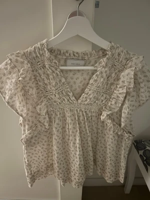 Blommig volangblus från Neo Noir - Supersöt blus från Neo Noir i ljusbeige med småblommigt mönster. Blusen har v-ringning, volangdetaljer längs axlar och bröst samt smockad överdel. Tillverkad i ett lätt och luftigt tyg, perfekt för en romantisk look.