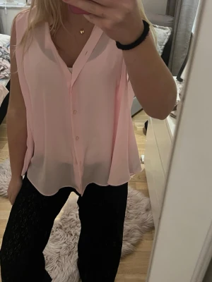 Ljusrosa ärmlös blus med knappar - Säljer en ljusrosa, flowy blus med knappar framtill och v-ringning. Blusen är ärmlös och har en lös passform som ger en avslappnad och luftig känsla. Perfekt att styla med både jeans och kjol. Materialet känns lätt och svalt.