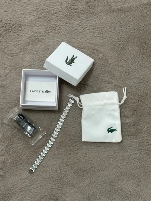 Silverfärgat armband från Lacoste - Snyggt silverfärgat armband från Lacoste med stilren och modern design. Armbandet har en länkstruktur i metall och kommer med original ask och tygpåse med Lacoste-loggan. Perfekt accessoar för dig som gillar klassisk men trendig stil.