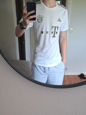 Vit Bayern München Adidas tröja - Snygg vit Bayern München t-shirt från Adidas med guldiga detaljer och klubbmärke på bröstet. Tröjan har korta ärmar, rund hals och tre guldiga ränder på axlarna. Perfekt för fotbollsfans som vill ha en stilren supportertröja.