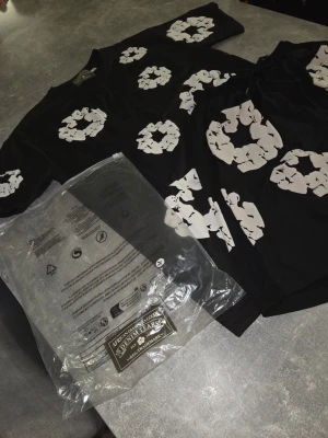 Svart t-shirt med vita tryck från Denim Tears - Säljer en svart t-shirt från Denim Tears med stora vita blommönster tryckta över hela tröjan. T-shirten har rund hals och korta ärmar. Materialet är mjuk bomull som känns skön mot huden. Perfekt för dig som vill sticka ut med en unik och trendig look.