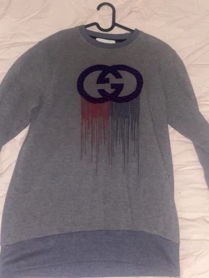 Grå sweatshirt med tryck - Säljer en grå sweatshirt med ett stort tryck framtill i lila och rött som smälter ner i ett coolt mönster. Tröjan har rund halsringning, långa ärmar och ribbade muddar vid ärmslut och nederkant. Perfekt för dig som gillar streetwear och vill sticka ut.