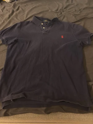 Marinblå pikétröja från Polo Ralph Lauren - Klassisk marinblå pikétröja från Polo Ralph Lauren med två knappar i halsen och den ikoniska röda logon broderad på bröstet. Tröjan har korta ärmar och är tillverkad i mjuk bomull som känns skön mot huden.