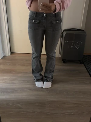 Gråa bootcut jeans med fickdetaljer - Säljer ett par gråa jeans bootcut med snygga fickdetaljer framtill. Jeansen har normal midja och två knappar i linningen. Materialet ser ut att vara klassisk denim, perfekt för en avslappnad och trendig look. Finns små blå färgfläckar 