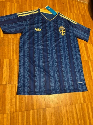 Blå Sverige fotbollströja Adidas - Säljer en blå fotbollströja med Sveriges landslagsemblem på bröstet. Tröjan har gula detaljer,  och ett unikt mönster över hela tyget. Perfekt för dig som vill stötta Sverige med stil.