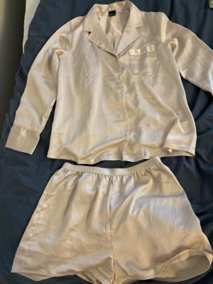 Beige satinpyjamas från Gina Tricot - Snygg tvådelad pyjamas från Gina Tricot i beige satin. Setet består av en långärmad skjorta med krage, bröstficka och knappar framtill samt matchande shorts med resår i midjan. 
