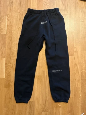 Svarta Essentials mjukisbyxor - Svarta mjukisbyxor från Essentials med resår i midjan och benslut, samt dragsko i midjan. Byxorna har en enkel design med Essentials-logga på ena benet och är tillverkade i mjukt material för maximal komfort.