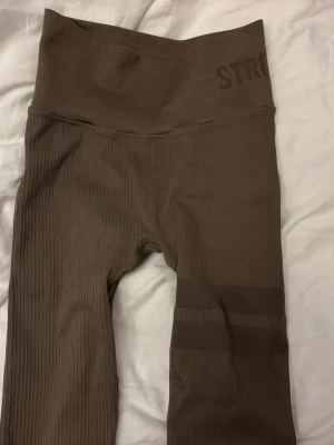 Bruna ribbade leggings från Stronger - Säljer ett par bruna ribbade leggings från Stronger med hög midja och bred linning. De har diskreta ränder på ena benet och är tillverkade i ett stretchigt material som sitter skönt på kroppen. Perfekta för träning eller chill dagar. Tycker att de passar S med!