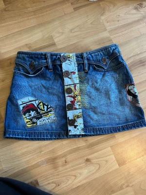 Kort jeanskjol med serietidningsmönster - Säljer en unik kort jeanskjol med serietidningsinspirerade detaljer och knappar framtill. Kjolen har blå denim som bas och färgglada tryck i pop art-stil på både knappslå och ficka. Perfekt för dig som vill sticka ut med en kreativ och cool look.