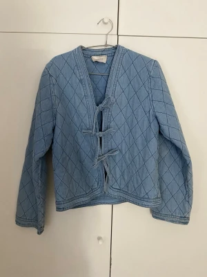 Ljusblå quiltad jeansjacka - Unik ljusblå jeansjacka med quiltat mönster och knytning framtill. Jackan har lång ärm, rak passform och V-ringad hals. Perfekt för dig som vill ha en annorlunda twist på den klassiska jeansjackan.