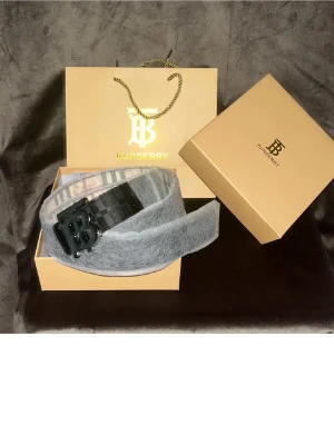 Svart Burberry bälte - Säljer ett grått bälte från Burberry med klassiskt rutigt mönster på insidan och ett svart BB-logga spänne. Bältet har en mjuk yta och känns riktigt lyxigt. Perfekt accessoar för att lyfta din outfit.