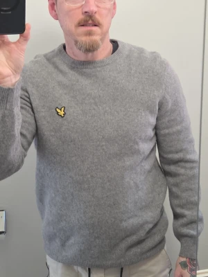 Grå stickad tröja från Lyle & Scott - Mjuk och stilren grå stickad tröja från Lyle & Scott med rund halsringning och den klassiska gula loggan på bröstet. Tröjan har långa ärmar och ribbade muddar vid ärmslut och nederkant. Perfekt för lager på lager och enkel att matcha.