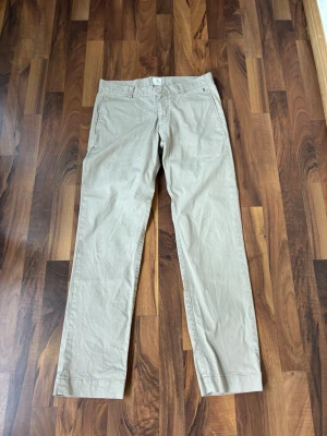 Beige chinos från Boomerang - Snygga beige chinos från Boomerang med rak passform och klassisk design. Byxorna har diskreta fickor fram och bak samt ett stilrent innerfoder med blommönster i midjan. Perfekta för en clean och avslappnad look.