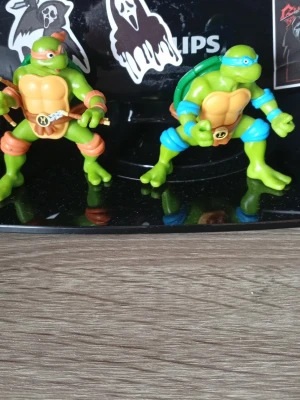 Teenage Mutant Ninja Turtles figurer - Två actionfigurer föreställande Teenage Mutant Ninja Turtles. Den ena har orange mask och bälten, den andra har blå mask och bälten. Båda är gröna med skal på ryggen och detaljer i plast. Perfekta för samlare eller som leksaker.