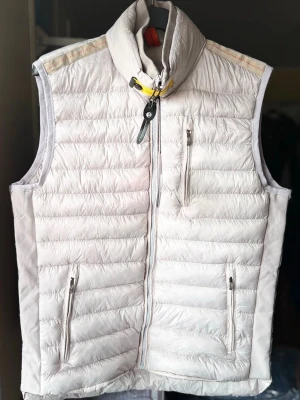 Vit dunväst från Parajumpers  - Snygg och lätt vadderad vit dunväst med hög krage och dragkedja framtill. Västen har tre fickor med dragkedja och en cool detalj med gul rem vid halsen. Perfekt för lager-på-lager och ger en clean look.