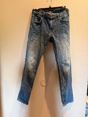 Blå raka jeans från Lee - Säljer ett par klassiska blå jeans från Lee med raka ben och fem fickor. Jeansen har snygga slitningar och kontrastsömmar, samt Lee-logga på knapp och etikett. Perfekta till en avslappnad stil och tillverkade i slitstark denim.