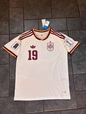 Vit Spanien fotbollströja Adidas - Vit Spanien fotbollströja med vinröda detaljer och nummer 19. Tröjan har Lamine Yamal tryckt på ryggen, broderat landslagsemblem och Adidas-logga framtill. Ränder på axlarna och patchar på ärmarna. Tillverkad i lätt och ventilerande material.