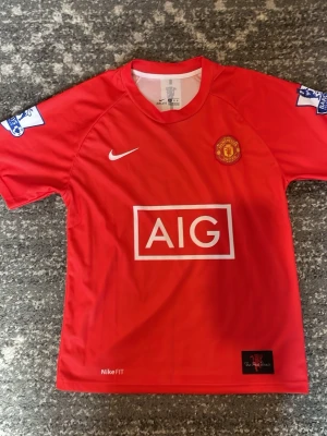 Manchester United matchtröja Nike röd - Klassisk röd Manchester United matchtröja från Nike med AIG som sponsor på bröstet. Tröjan har klubbmärke på vänster bröst, Nike-logga på höger och Premier League-patch på ärmarna. Tillverkad i funktionsmaterial med korta ärmar och rund hals.Ronaldo på ryggen nummer 7. Storlek S