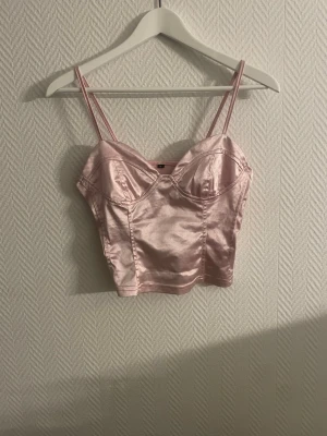 Rosa satin korsett-topp - Snygg ljusrosa korsett-topp i satin med smala axelband och croppad passform. Toppen har en glansig finish och markerade kupor framtill för en trendig look. Perfekt att styla med jeans eller kjol för en cool outfit.