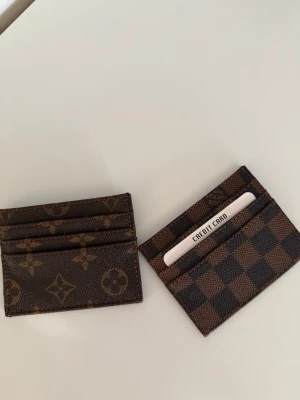 Bruna kortfodral - Två stilrena kortfodral i brunt läder. Ena fodralet har klassiskt monogrammönster och det andra har rutigt Damier-mönster. Båda har flera kortfack och är perfekta för att hålla ordning på dina kort. Snygga accessoarer med lyxig känsla. Säljer båda tillsammans. Annars kan gör göra 1 för 150kr