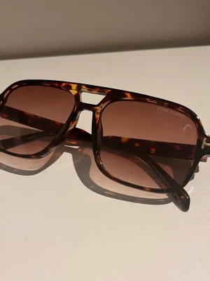 Tom Ford bruna solglasögon - Snygga solglasögon från Tom Ford med brunmelerad båge i klassisk pilotmodell. Glasen är bruna och bågen har ett coolt mönster som ger en exklusiv känsla. Perfekta för att lyfta din stil under soliga dagar.