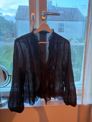 Svart transparent volangblus - Snygg svart blus i transparent material med volangdetaljer längs knäppningen och kragen. Blusen har långa ärmar och ett lätt, luftigt tyg som ger en cool och trendig look. Perfekt för att lyfta vilken outfit som helst. Andvänd 1 gång 🥰perfekt till ett linne under eller en snygg BH 😍 pris kan diskuteras 