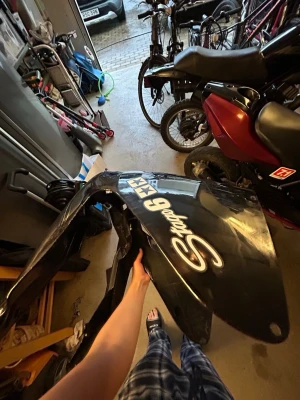 Svart kåpa med S mopeddekal - Säljer en svart plastkåpa till moped med vit text 'Stage 6' och dekaler. Kåpan har en böjd form och passar till styling eller reservdel. Perfekt om du vill fixa till din moppe och ge den en personlig touch.