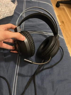 Svarta gaminghörlurar med mikrofon - Säljer ett par svarta over-ear gaminghörlurar med inbyggd mikrofon och vadderad huvudbygel. Hörlurarna har en cool ram-logga på kåporna, mjuka öronkuddar i konstläder och slitstark flätad kabel med både ljud- och mikrofonkontakt. Perfekta för gaming eller videosamtal.