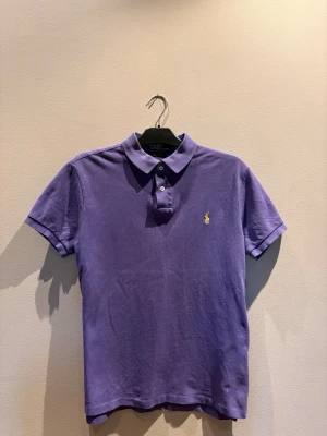 Lila pikétröja från Ralph Lauren - Klassisk lila pikétröja från Ralph Lauren med korta ärmar och krage. Tröjan har två knappar framtill och den ikoniska broderade loggan i gult på bröstet. Perfekt till jeans eller shorts för en avslappnad men snygg look.