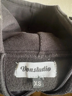 Svart hoodie med dragkedja Vonstudio XS - Svart hoodie från Vonstudio i storlek XS. Hoodien har dragkedja framtill, huva med snören och coolt tryck med text och ett lejon i vitt på bröstet. Perfekt för dig som gillar streetwear och vill ha en unik look.