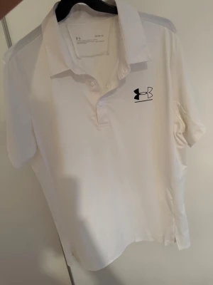 Vit pikétröja från Under Armour - Snygg vit pikétröja från Under Armour med klassisk krage och knappar framtill. Tröjan har korta ärmar och är tillverkad i ett lätt och stretchigt material som känns skönt mot huden. Perfekt för en sportig och clean look.
